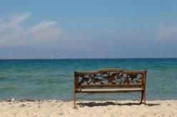 Das Bild zeigt eine Bank aus Holz vor dem Strand und dem Meer. Das Bild steht für Urlaub, Urlaubsgeld und Urlaubsabgeltung 